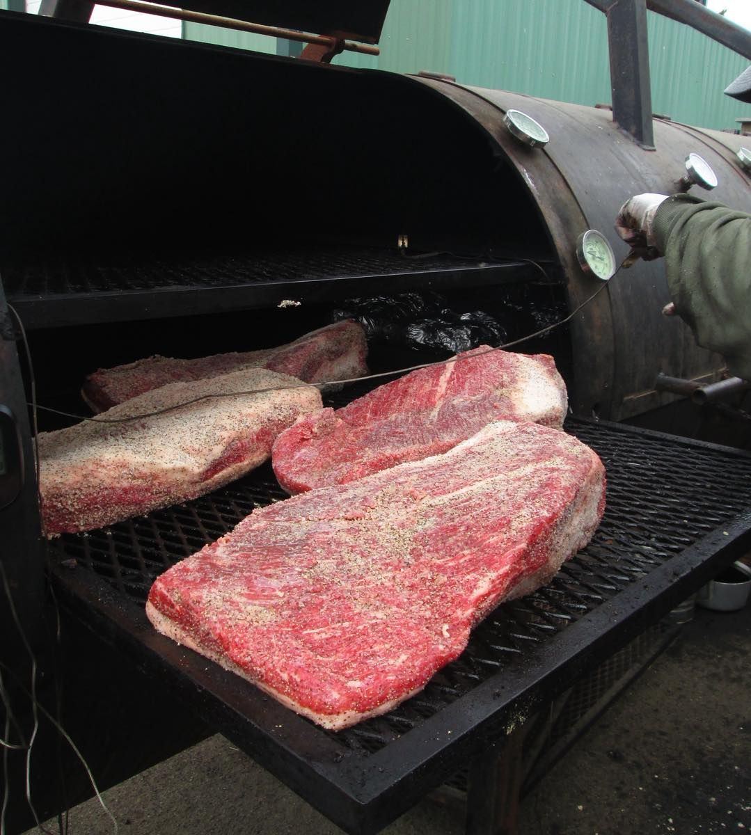 Seasoned whole briskets laid out on the grate of a big offset smoker, a gloved hand at the lid