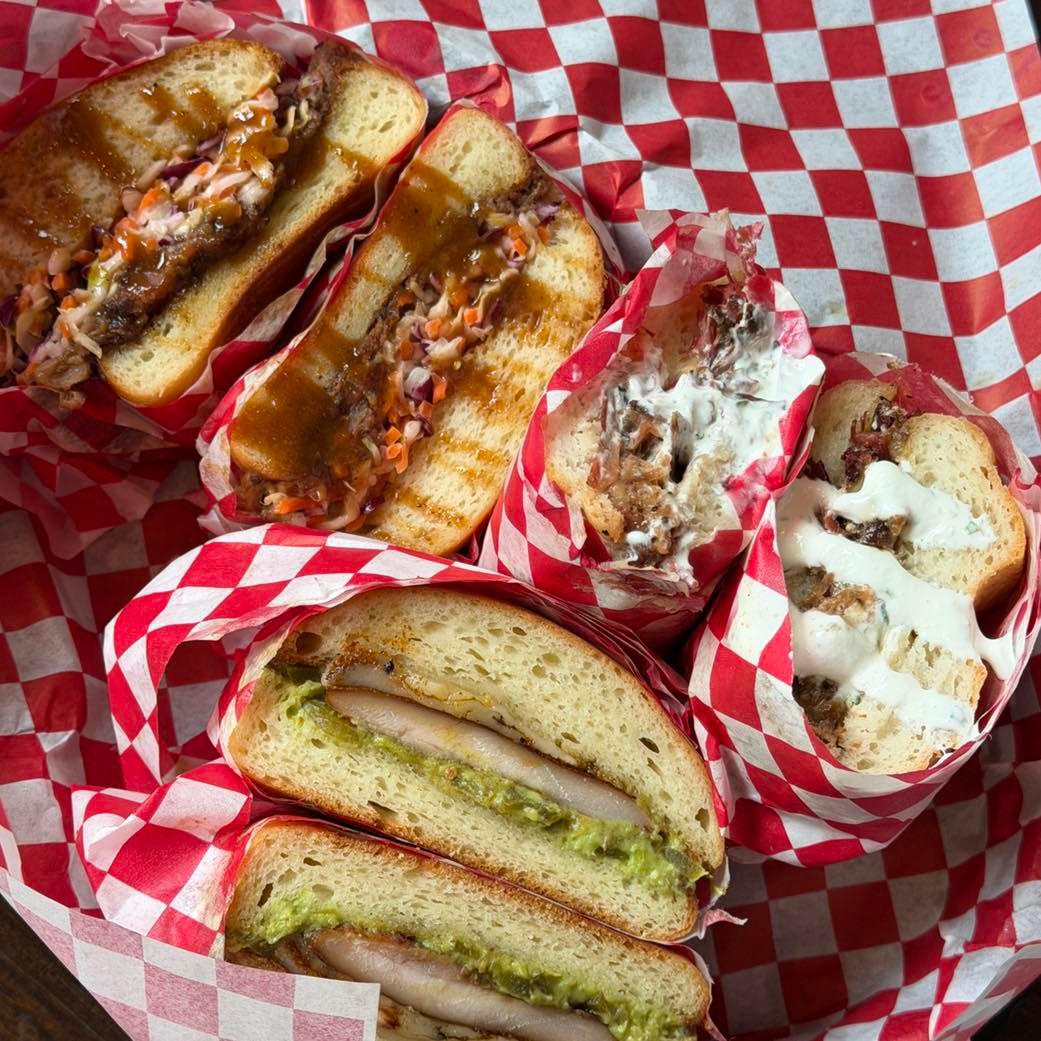 Overhead view of halved smoked sandwiches in red-and-white checkered paper, some with slaw and sauce, others with avocado crema