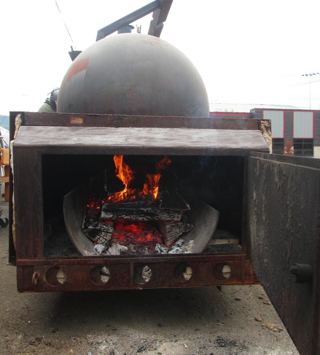 The smoker’s firebox door open in daylight, split logs burning over glowing coals